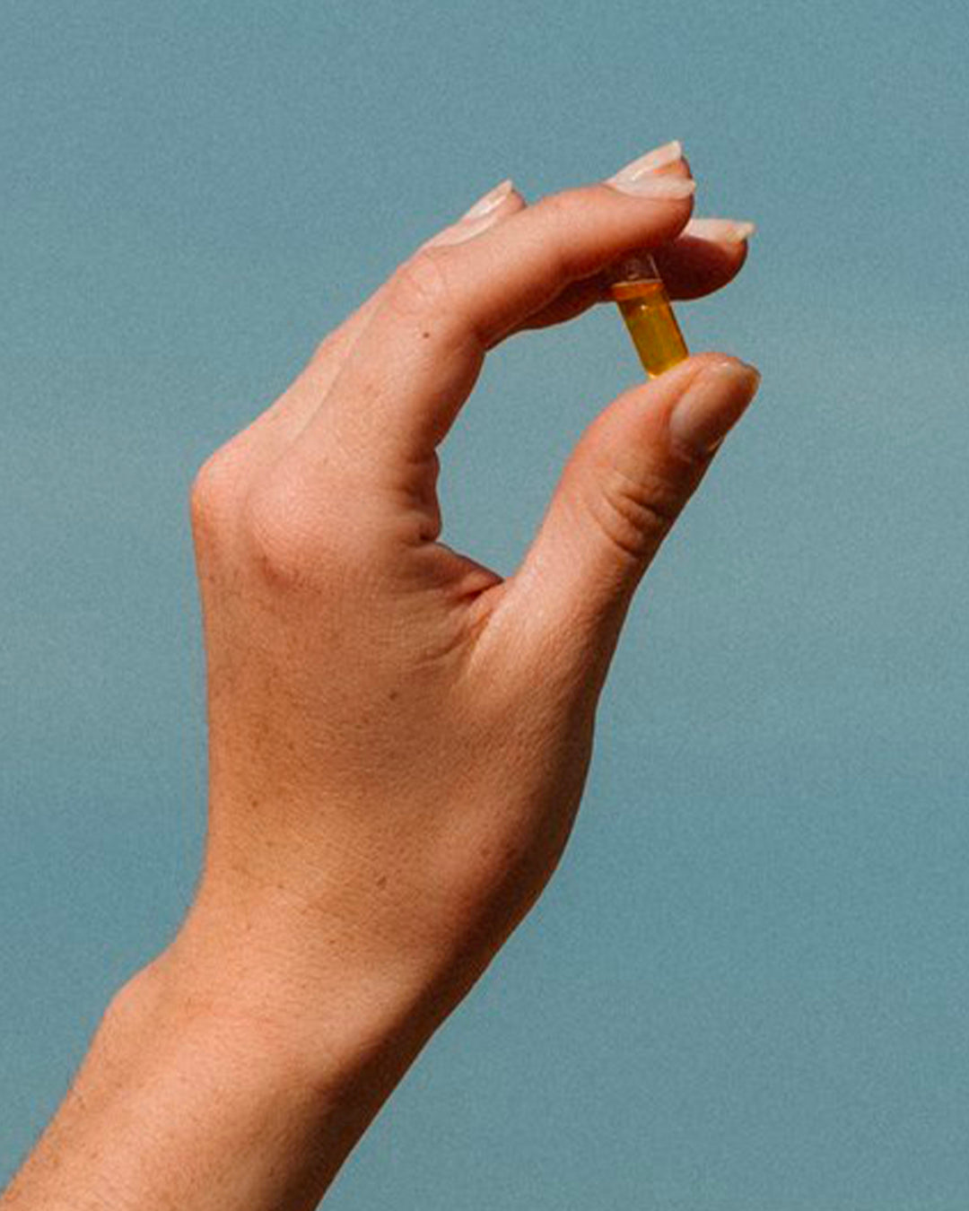 Atelier Nubio Plump Up My Skin, S'il Vous Plait golden capsule held between finger and thumb against a blue sky