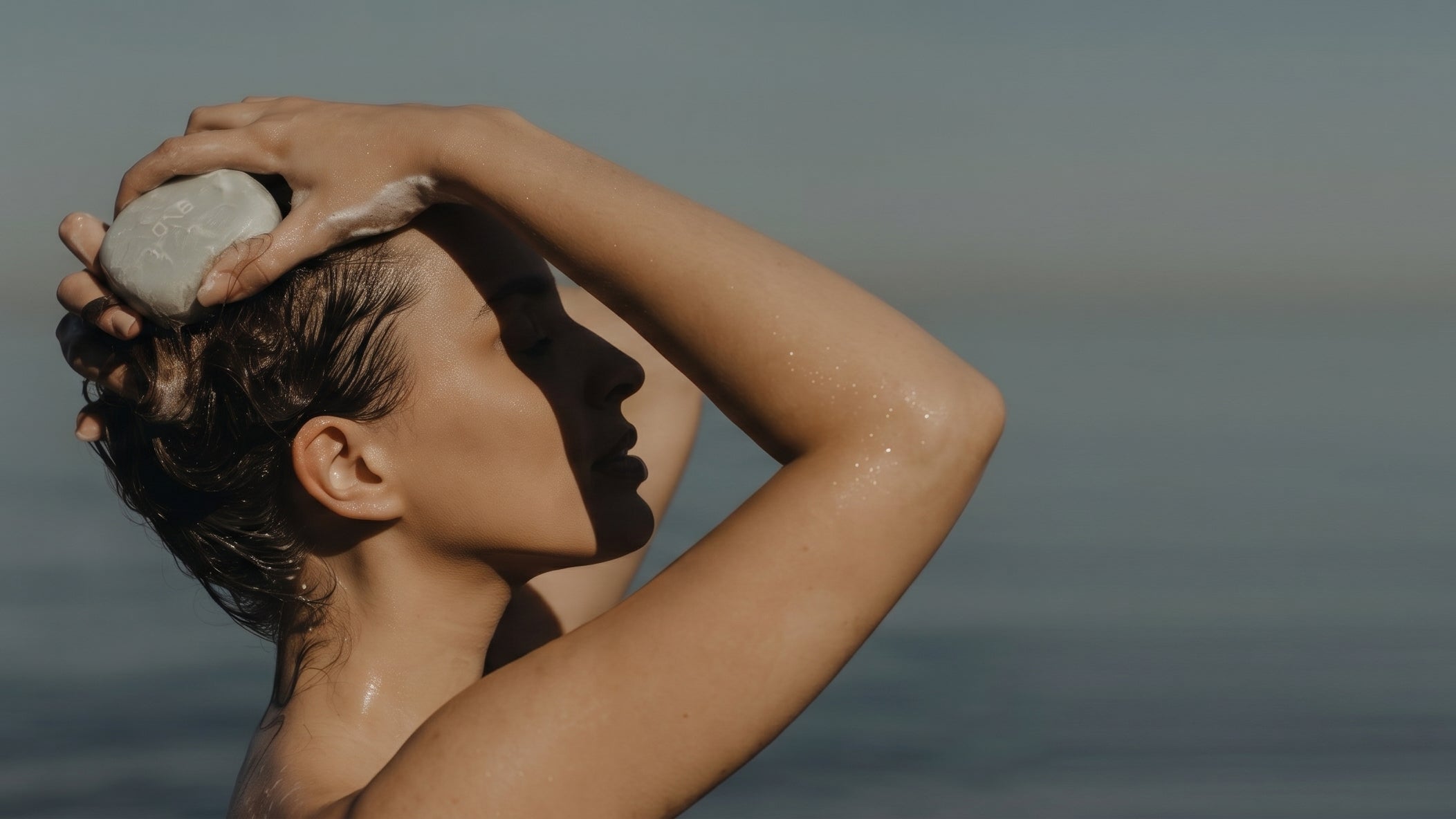 Model lathering a Beauty Disrupted solid shampoo bar into her hair by the sea