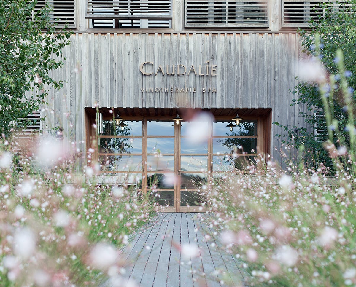 Entrance of the Les Sources de Caudalie Vinotherapie Spa, timber facade with carved Caudalie signage behind wildflowers