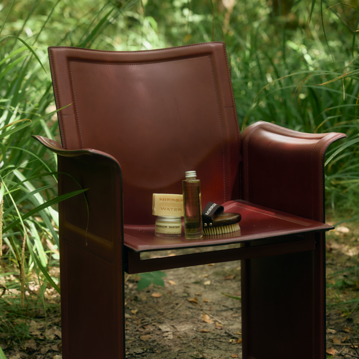 MIRROR WATER Shea Butter Brick with SMOOTH Body Oil and BUFF brush on a leather chair among ferns