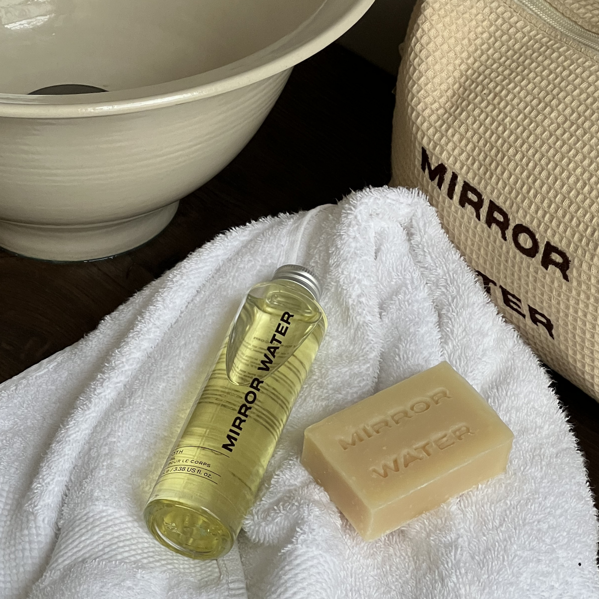 MIRROR WATER Shea Butter Brick and SMOOTH Body Oil bottle on a white towel beside a ceramic basin