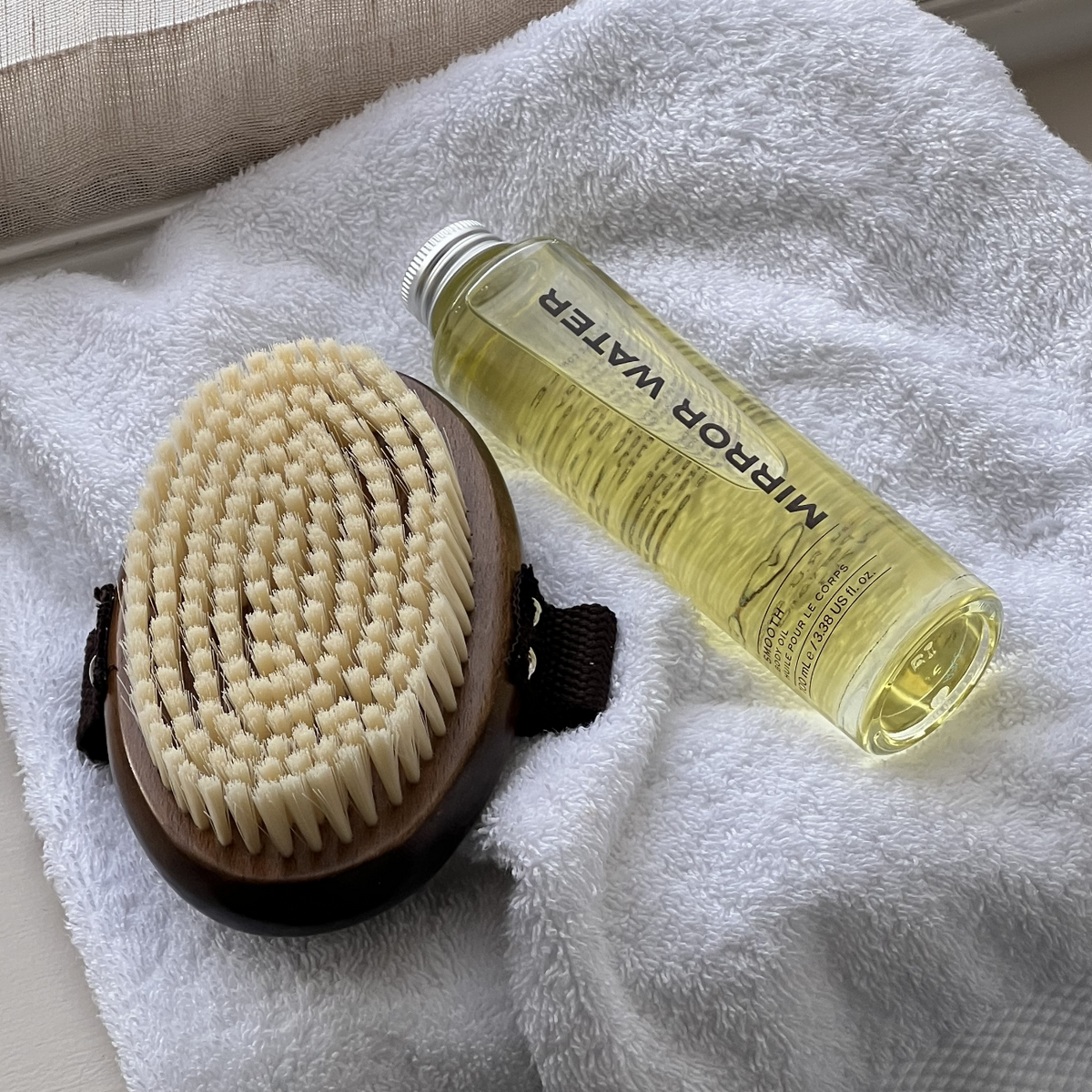 MIRROR WATER SMOOTH Body Oil bottle beside the BUFF Body Dry Brush on a white towel