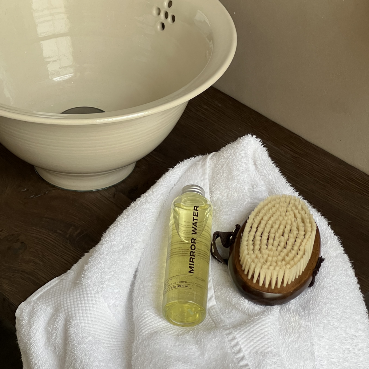 MIRROR WATER Ultra-Soft BUFF Body Dry Brush on a white towel beside a SMOOTH Body Oil bottle and a ceramic basin