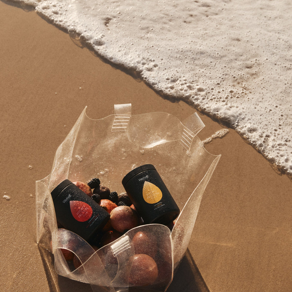 WelleCo The HydroProtein Elixir two jars in a clear bag with blackberries and passionfruit on a beach